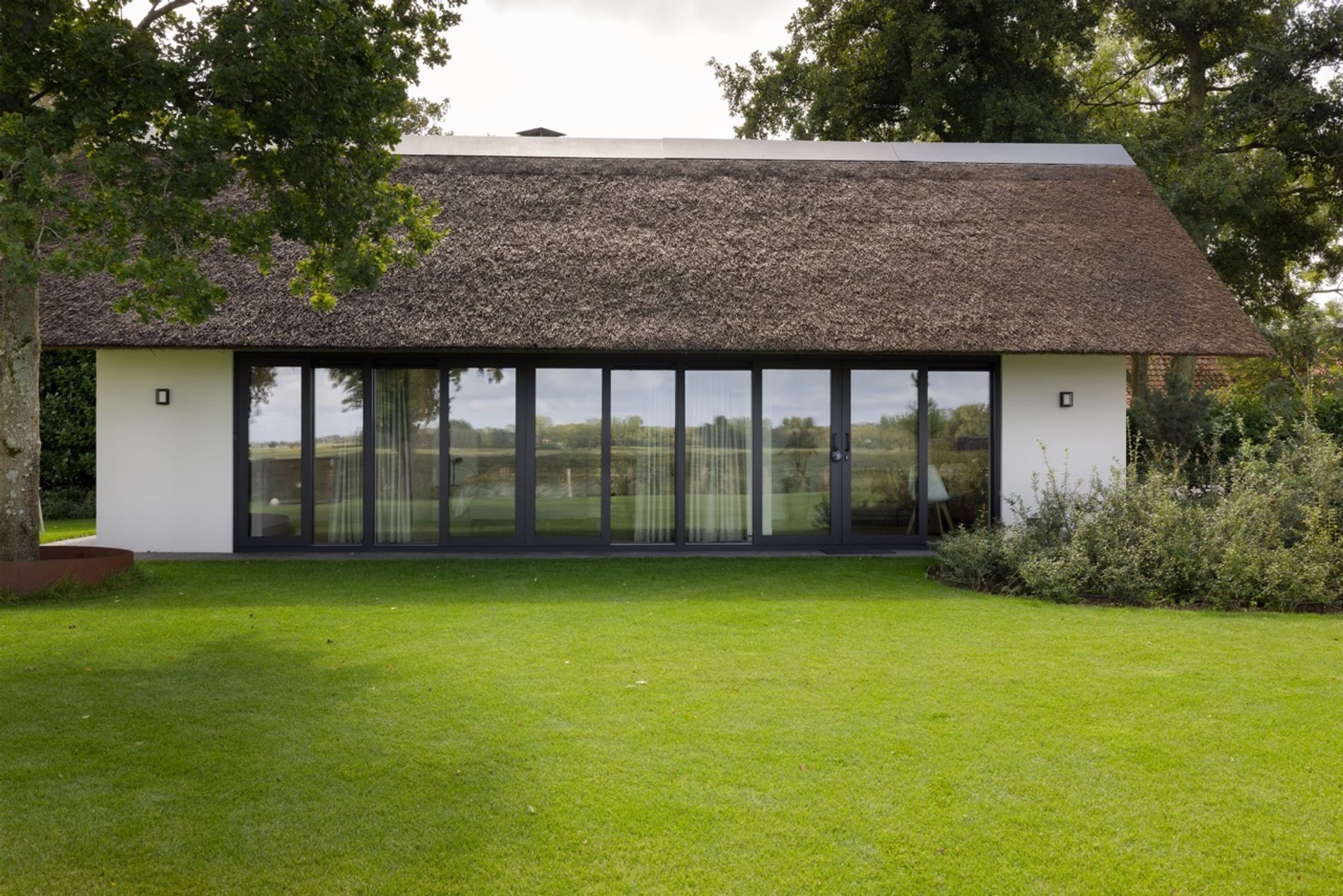 Die acht Meter breite Glasfassade des Gästehauses spiegelt die Polderlandschaft unter der tiefen Linie des Reetdachs.